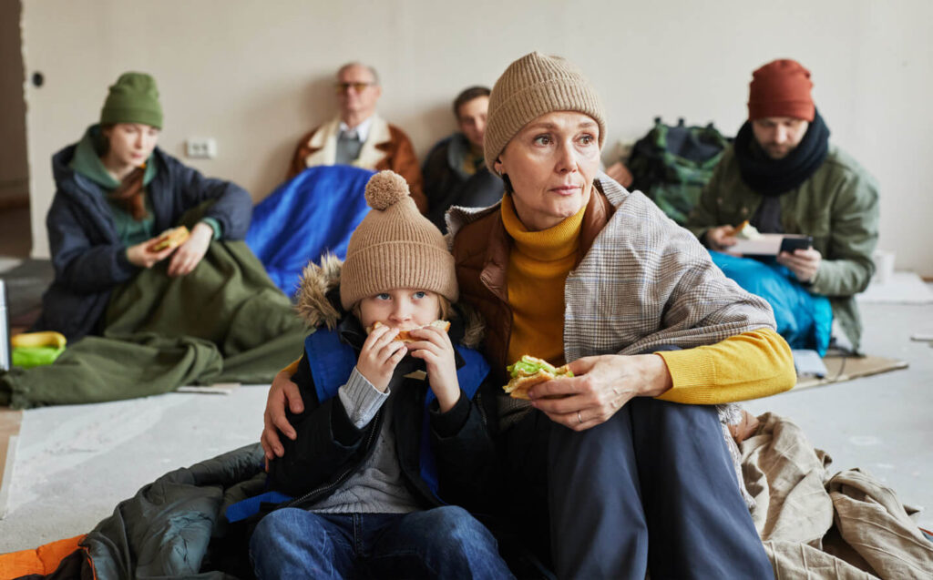 In a crowded post-disaster shelter, a woman and child sit on the floor surrounded by blankets, highlighting their difficult situation.